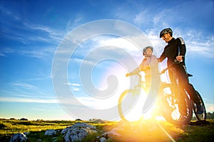 Cyclist couple with mountain bikes standing on the hill under the evening sky and enjoying bright sun at the sunset.