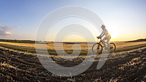 The cyclist rides a bike on the road near the field against the backdrop of the setting sun. Outdoor sports. Healthy lifestyle