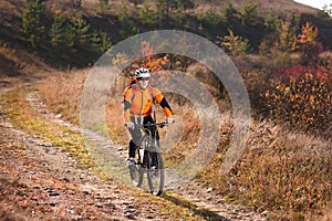 Cyclist in the sportive orange jacket riding a mountain bike along the trail in the countryside at sunset.