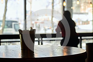 A cylindrical glass napkin on a brown table in a cafe. On a blurred background, the silhouette of a lonely girl at the bar counter