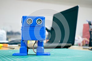 3D printed Funny blue robot on the background of devices and laptop. Robot model printed on automatic three dimensional 3d