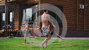 Dad circling his son in his arms such as on the carousel, and next to the mother and the second son playing with a hose