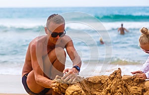 Dad and daughter make sand castle on the shore of sandy tropical beach in the background of bathing people