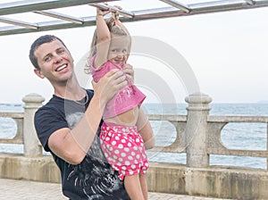 Dad and daughter playing promenade by the sea. young man, little girl hanging on the bar.