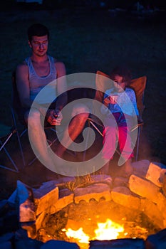 Dad and daughter sit at night by the fire in the open air in the summer in nature. Family camping trip, gatherings around the