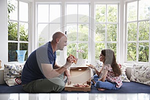 Dad and daughter sit on window seat at home sharing a pizza