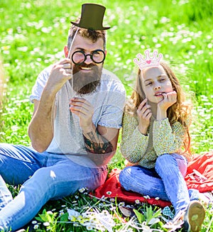 Dad and daughter sits on grass at grassplot, green background. Child and father posing with eyeglases, crown and top hat