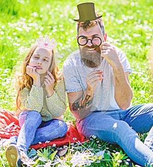 Dad and daughter sits on grass at grassplot, green background. Child and father posing with eyeglases, crown and top hat