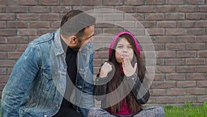 Dad and daughter are sitting, having fun and chewing bubble gum against the backdrop of grass and a brick wall. Concept