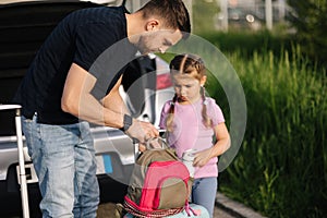 Dad and daughter are standing with their suitcases near a car with an open trunk. Happy little girl preparing to travel.