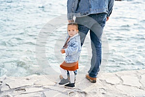 Dad in a denim jacket and jeans holds a little girl by the hand, standing on the rocks by the water. Girl turned to face