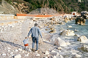 Dad in a denim jacket and jeans walks with a little girl holding her hand along a rocky pebble beach near the water