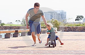 Dad has his back while he rides his bike. A happy toddler boy riding is toy motorbike outside while his father runs