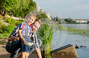 Dad helps the Little Boy to lower the paper boat to the water.