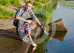 Dad helps the Little Boy to lower the paper boat to the water.