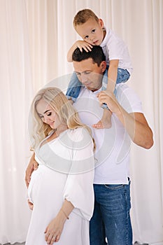 Dad hold his son on neck and hug his pregnat wife. Family look concept for studio photosession. Perents waiting for baby