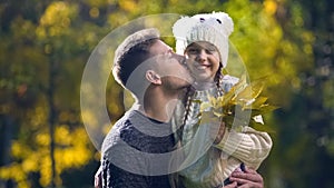 Dad holding daughter in funny hat and kissing, leisure together, autumn stroll