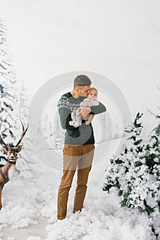 Dad holds a baby son in his arms in a winter forest style photo zone with a deer and dad kisses his son