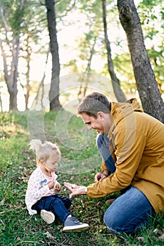 Dad holds out a tiny mushroom in the palm to a little girl sitting in the forest on the grass