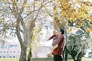 Dad kisses a little girl in his arms on the cheek while standing under a tree with yellow leaves