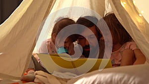 Dad and little daughters reading book in teepee tent