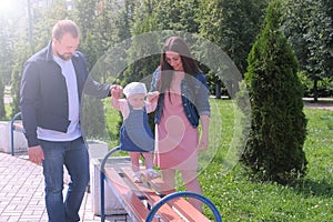 Dad and mom holding hands baby girl going by bench in park at summer.