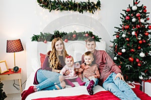 The dad, mom hug little son and daughter on bed in bedroom near Christmas tree. Happy New Year and Merry Christmas