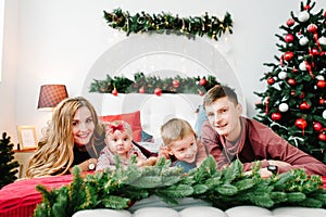 The dad, mom hug little son and daughter on bed in bedroom near Christmas tree. Happy New Year and Merry Christmas