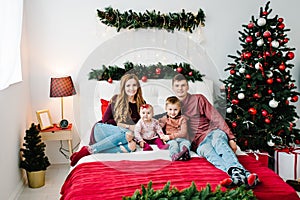 The dad, mom hug little son and daughter on bed in bedroom near Christmas tree. Happy New Year and Merry Christmas. Christmas