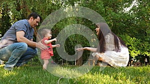 Dad and Mom learn to walk little daughter in park on green grass. Mom kisses daughter on the cheek and laughs with dad.