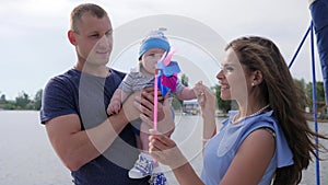 Dad and mom play with son at lake, woman entertains baby with help pinwheel on loch,