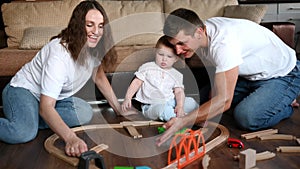 Dad and mom are relaxing in evening in a family circle, sitting on floor and playing iron wooden road with a happy