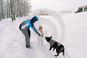 Dad plays with a black dog with a stick while standing with a little girl on a snowy village road