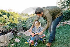 Dad pushes behind the wheel of a bike with a little girl pedaling on green grass