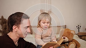 A dad reads his daughter a book on a bed in the room. Family home evening. A man and a girl play together, leafing through the pag