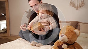 A dad reads his daughter a book on a bed in the room. Family home evening. A man and a girl play together, leafing through the pag