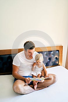 Dad is sitting on the bed with a little girl with a book in his hands