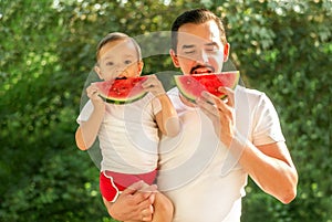 Dad and son eating and simultaneously biting slices of watermelon on picnic. Father is holding boy in arms