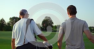 Dad son going fairway course. Two golfers take sport equipment clubs outdoors.