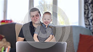 Dad and son are looking at the laptop. Video call with relatives. Dad and son wave `hello` into the laptop, talk with grandparents