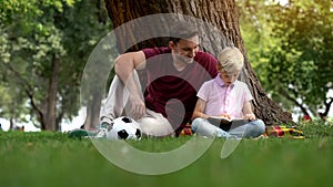 Dad and son reading book, sitting in park under tree, hobby, togetherness