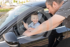 Dad is standing near the car in which the child is driving. The boy looks at the man and smiles