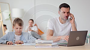 Dad talking on phone and son playing with gadget while sitting at table in home room spbi.