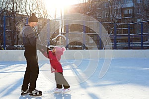 Dad teaches his little daughter to ice skate on a skating rink in the courtyard of multi-storey buildings in the city. Frosty