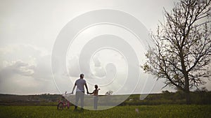 Dad is teaching daughter how to ride bicycle on the meadow at sunset time. Happy father rejoices that her daughter