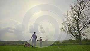 Dad is teaching daughter how to ride bicycle on the meadow at sunset time. Happy father rejoices that her daughter