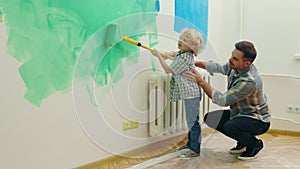 Dad teaching little handsome kid to paint wall using roller brush. Happy child boy helping father to renovate house