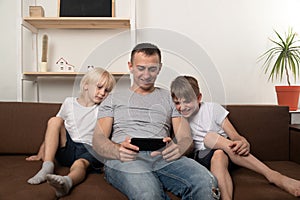 Dad and two sons are sitting on couch and watching video on phone. Family leisure