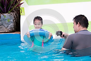 Dad is using camera to take video of daughter playing in pool with slide. Cute little girl hugged blue rubber ring.