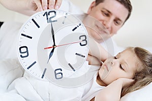 Dad wakes up little sleeping in bed girl,daughter. Big alarm,clock. Early morning before kindergaten, school. White pillow,